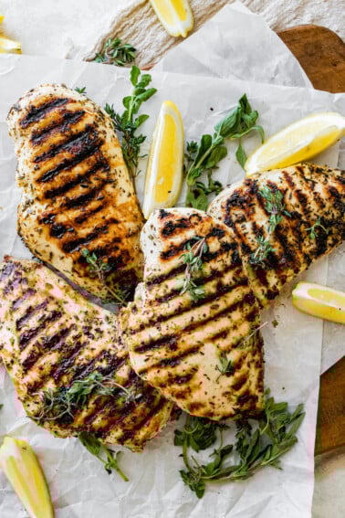 Four grilled chicken breasts on parchment paper with fresh herbs and lemon slices.